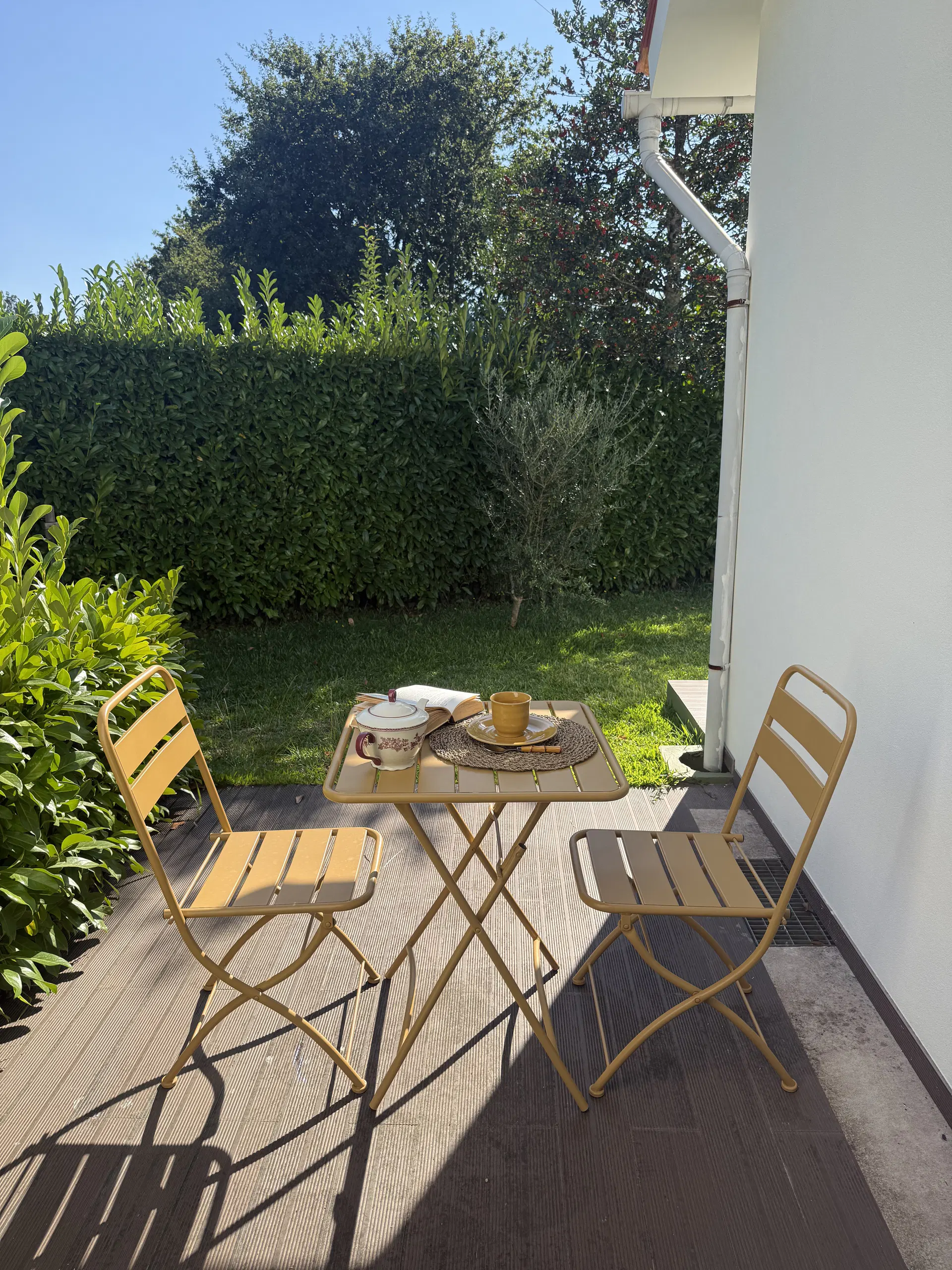 Mesa e cadeiras no pátio junto à casa, com o relvado e a sebe em frente.
