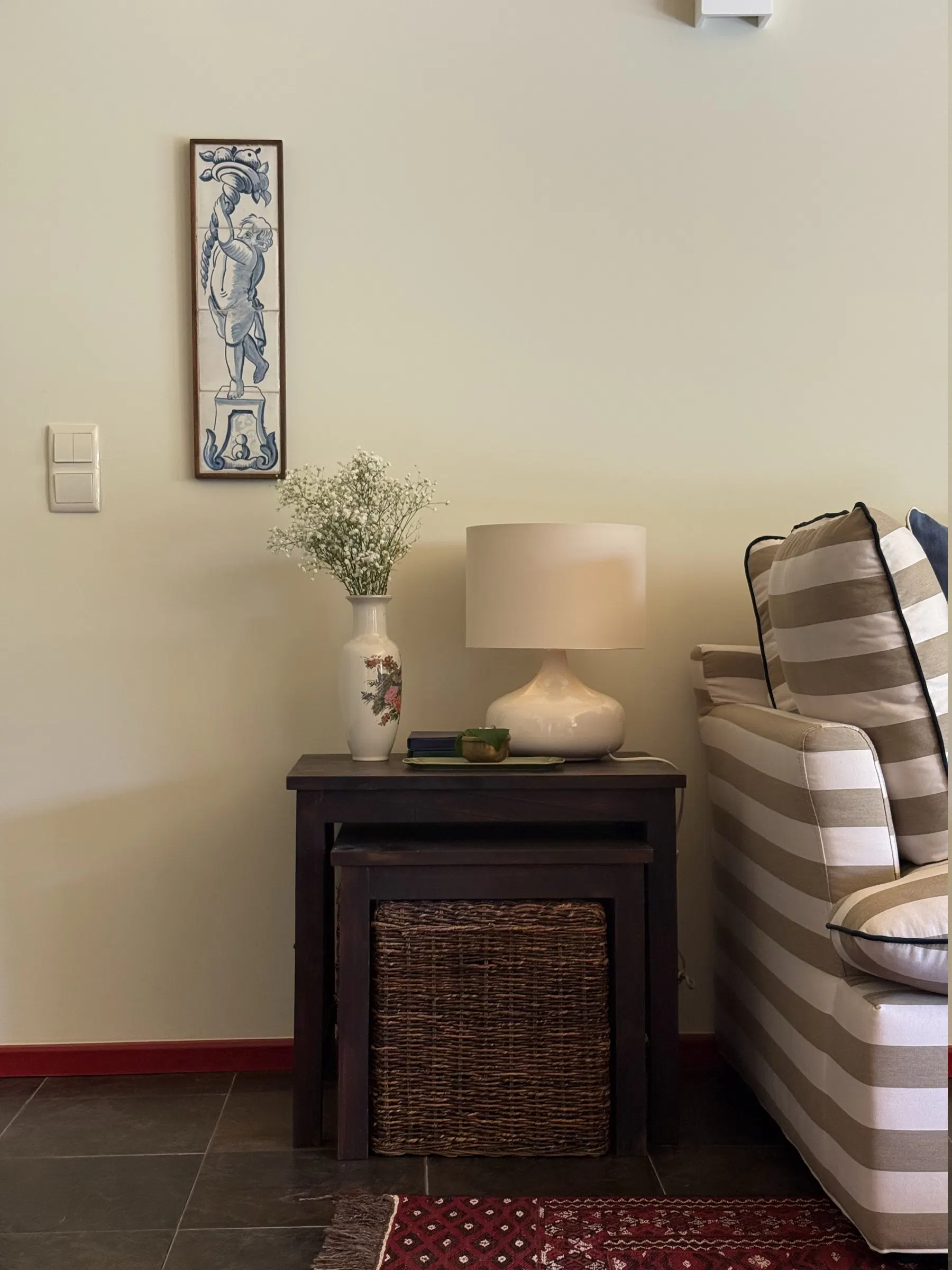 Pormenor da sala: painel de azulejos antigo, jarra de flores e candeeiro sobre mesas de encaixe.
