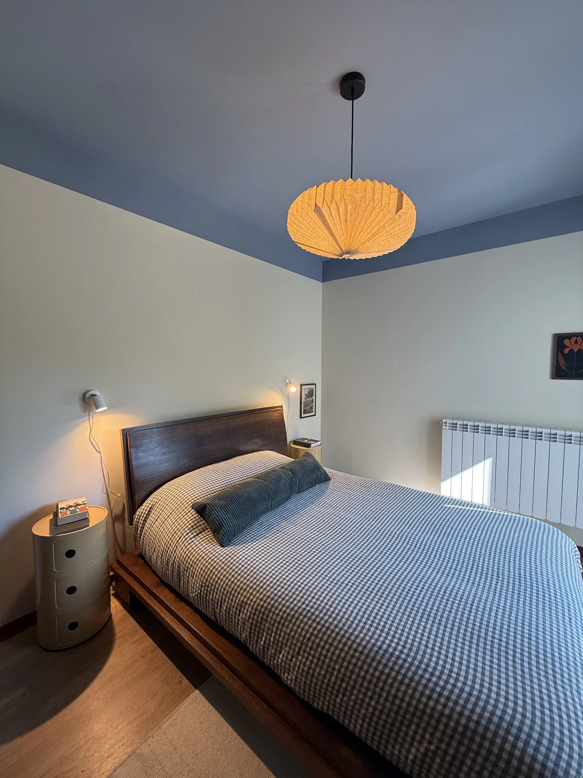 Quarto de casal com teto azul, cabeceira de madeira e candeeiro suspenso.