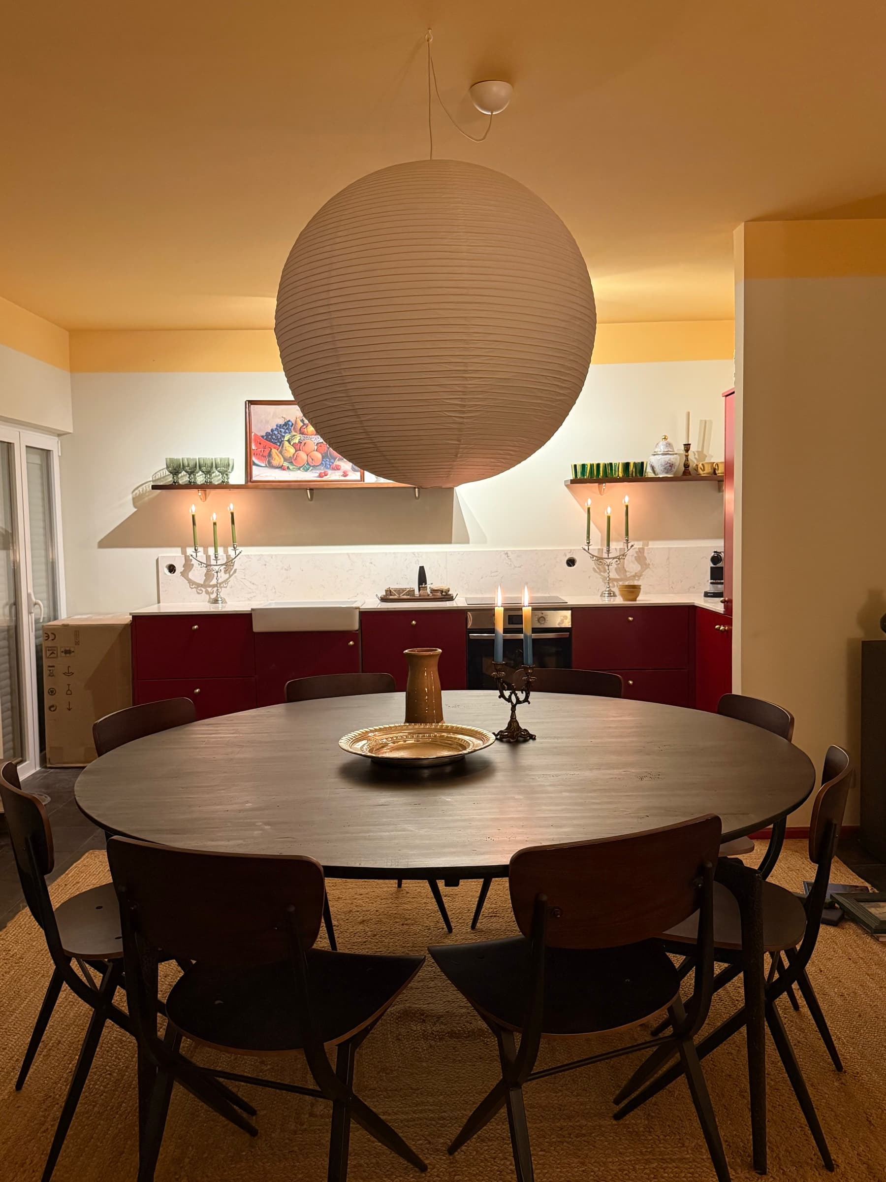 A mesa de jantar à noite, com velas em candelabros e a cozinha vermelha iluminada ao fundo.