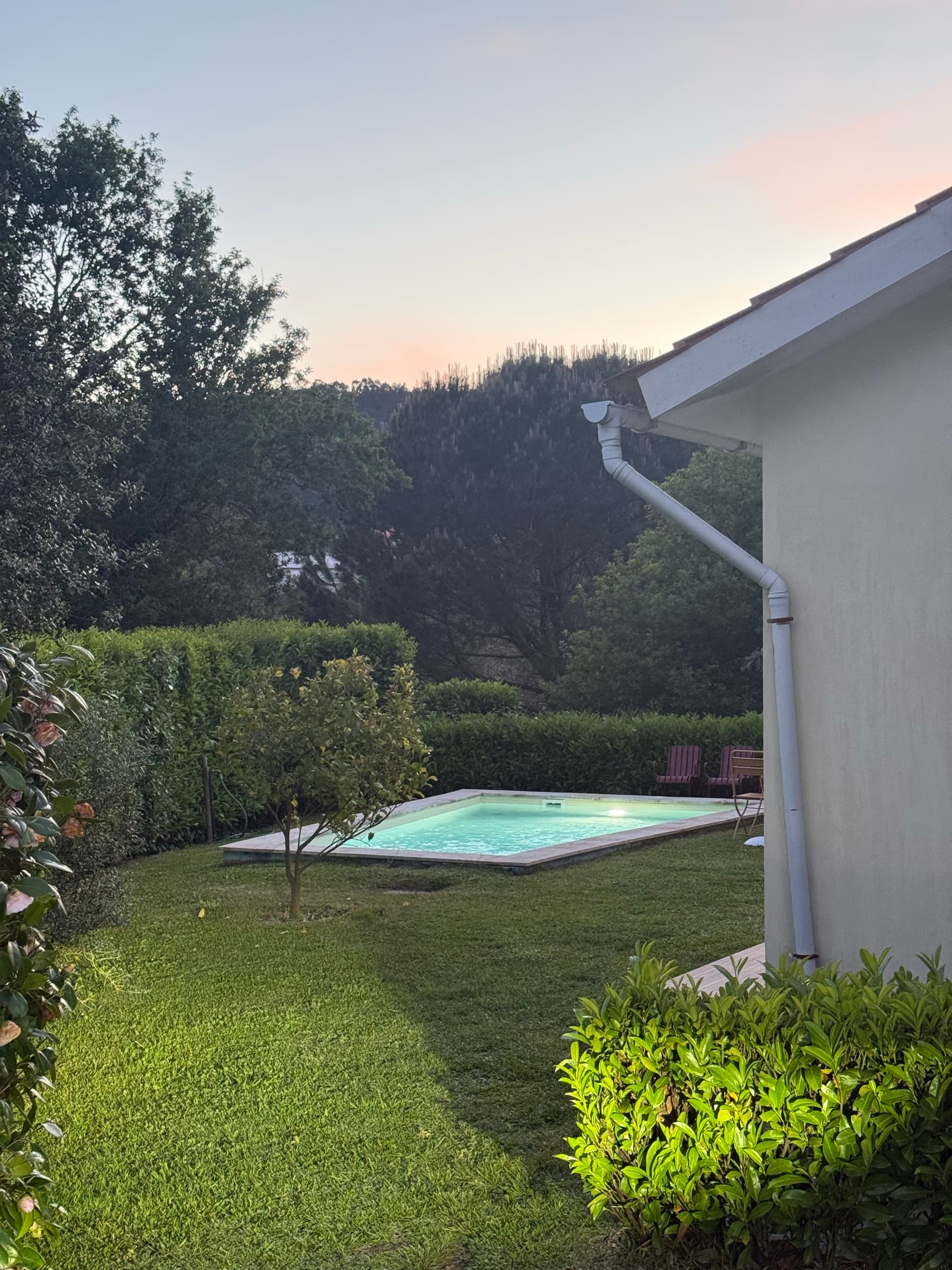 A piscina iluminada ao anoitecer, com o relvado, a sebe e o monte a escurecer atrás.