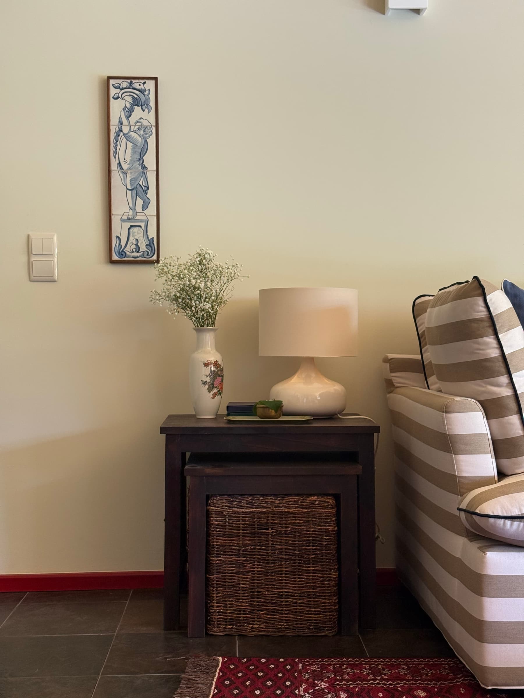 Pormenor da sala: painel de azulejos antigo, jarra de flores e candeeiro sobre mesas de encaixe.