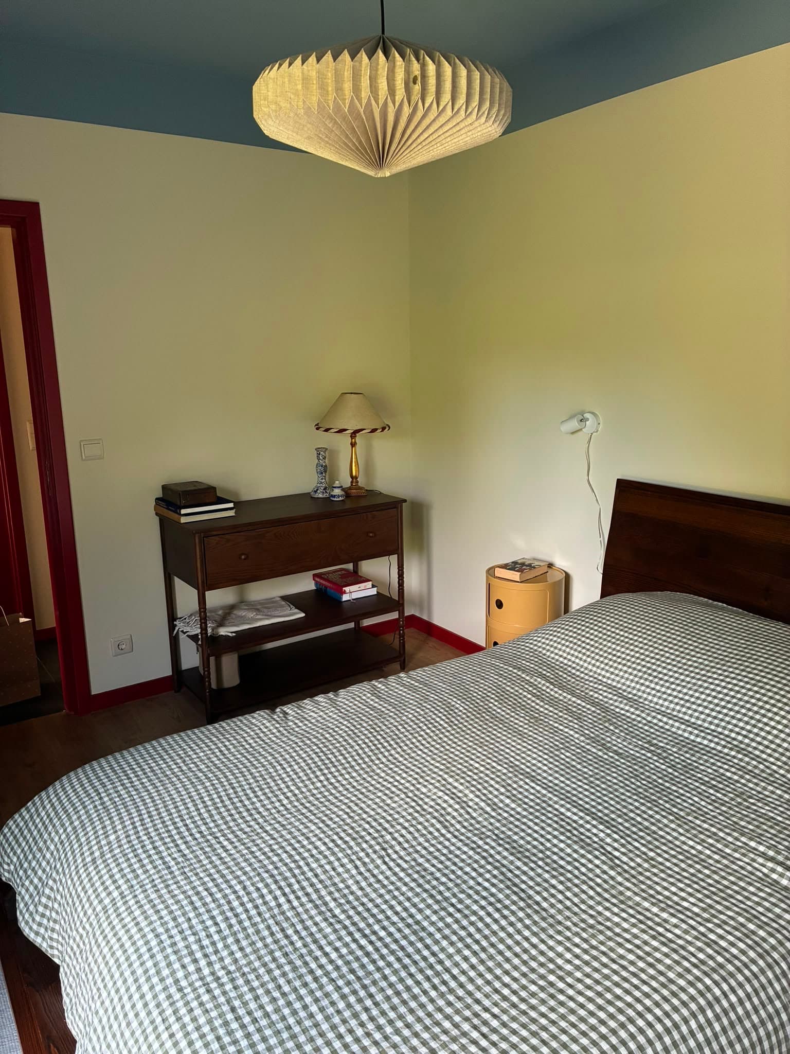 Quarto de casal com o teto pintado de azul, edredão aos quadrados e aparador de madeira escura.