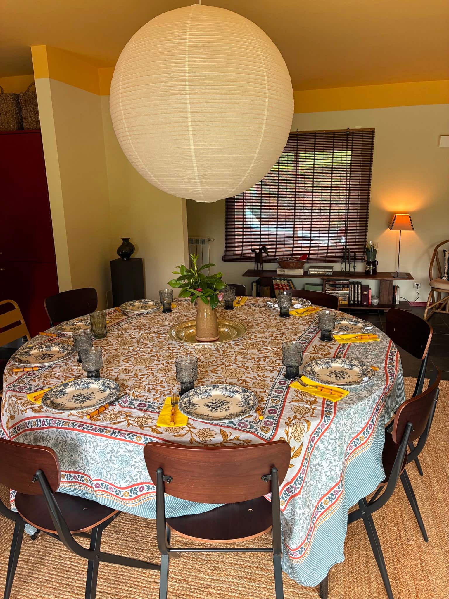 A mesa de jantar redonda posta com toalha estampada e loiça antiga, sob um candeeiro de papel.