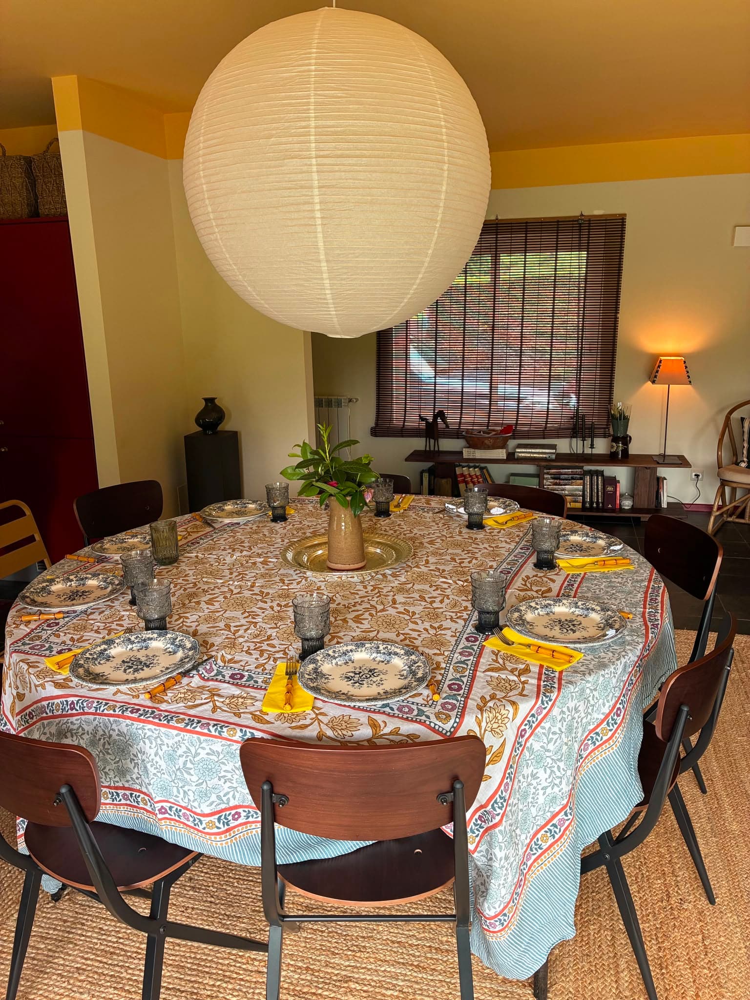 A mesa de jantar redonda posta com toalha estampada e loiça antiga, sob um candeeiro de papel.