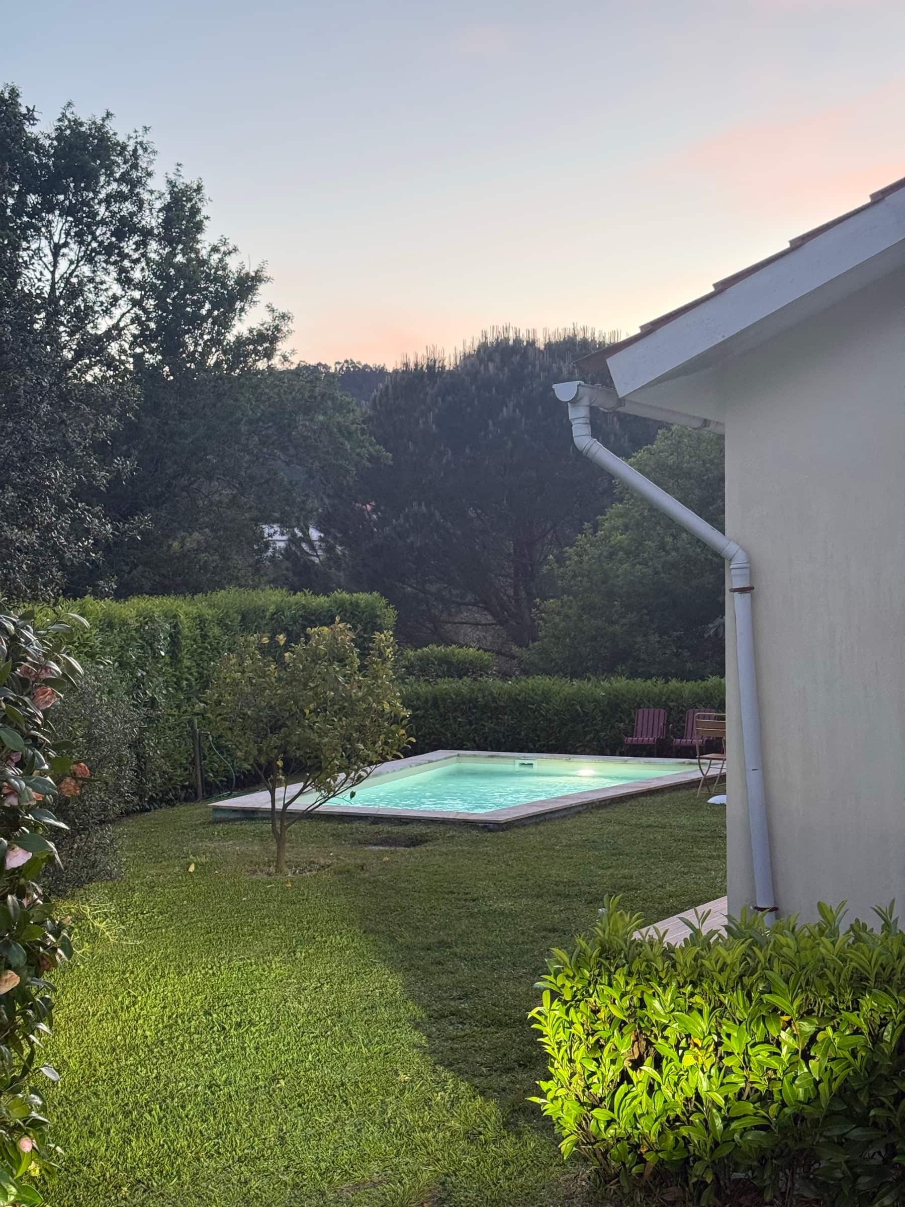 A piscina iluminada ao anoitecer, com o relvado, a sebe e o monte a escurecer atrás.