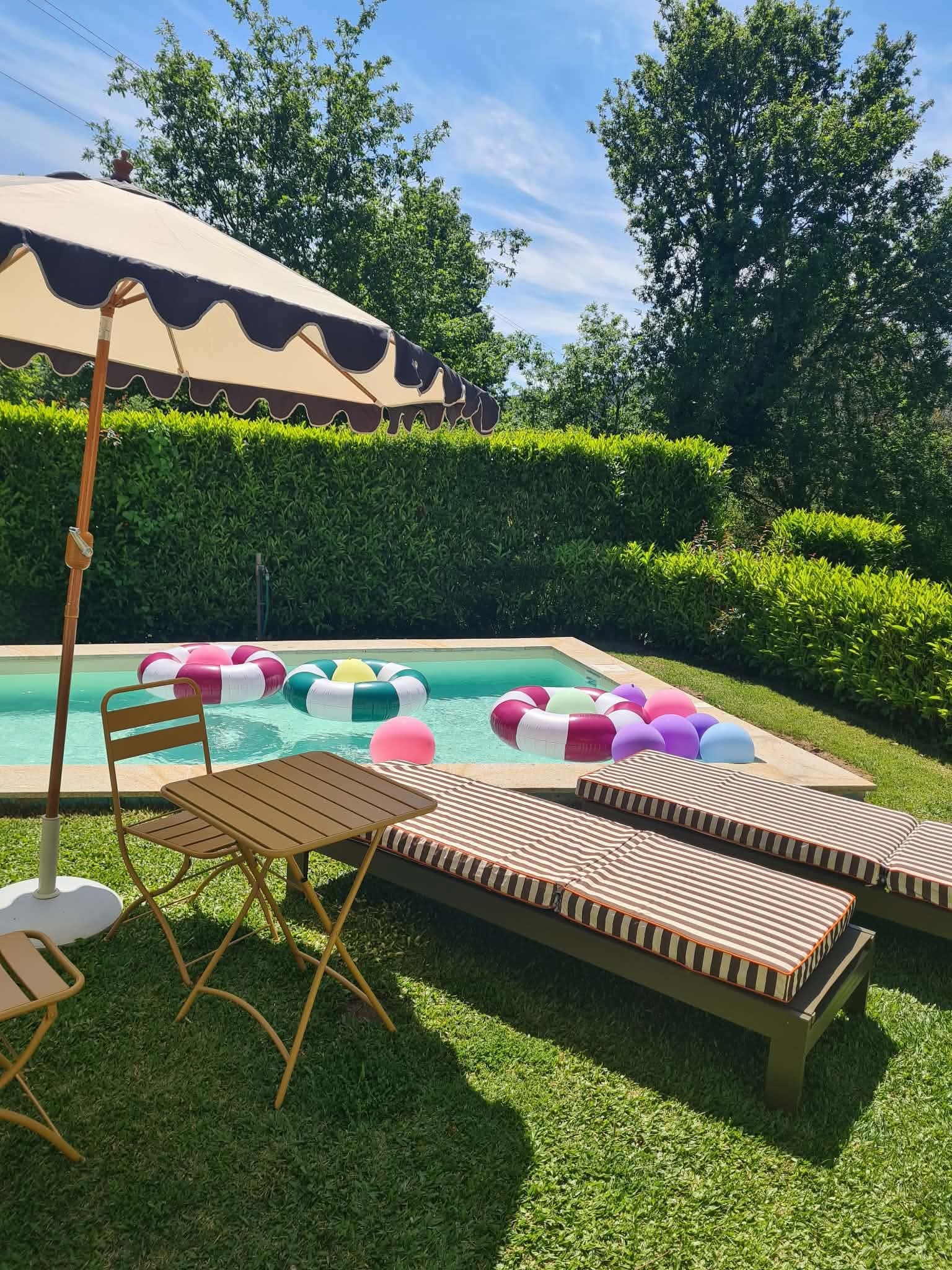 Espreguiçadeiras às riscas e guarda-sol de abas onduladas junto à piscina, com boias coloridas na água.